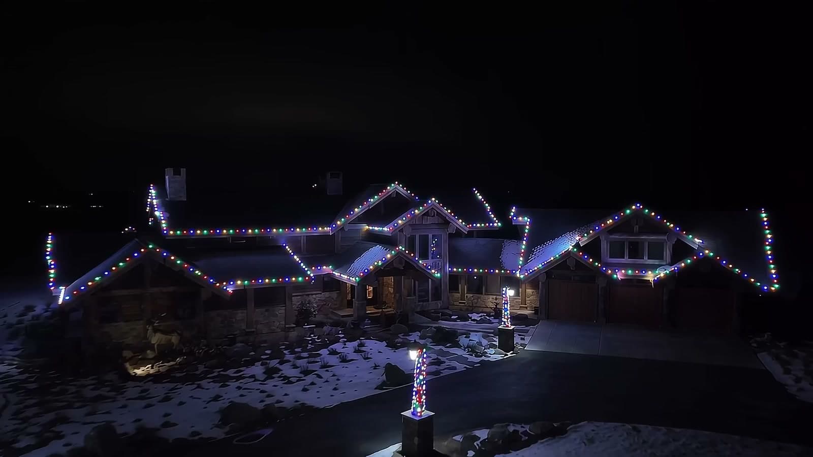 Aerial view at night of a large timber home outlined in multicolor roofline lights, with wrapped lamp posts along a snowy driveway
