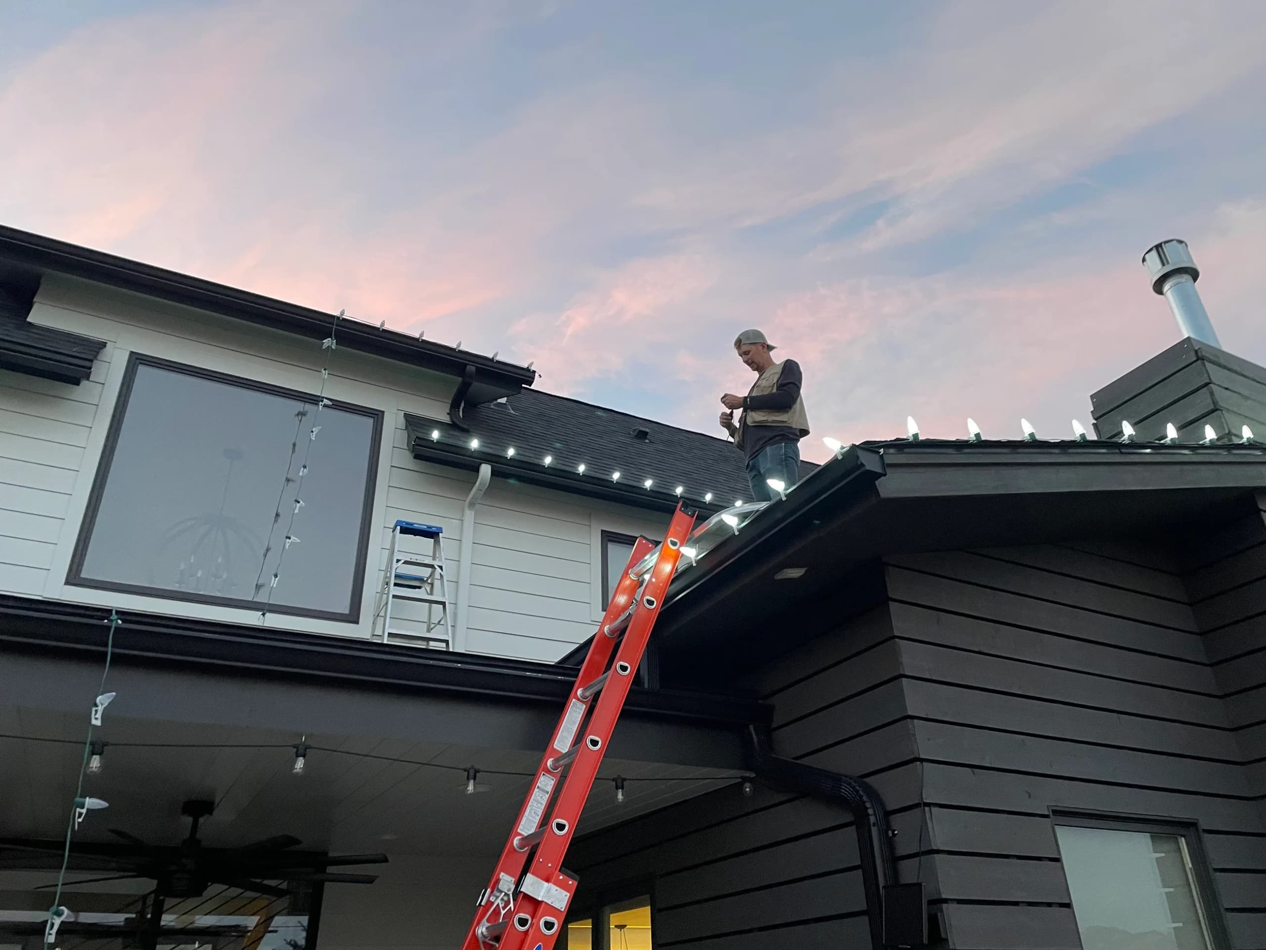 Installer working on a roof beside a ladder, clipping lights along the roofline
