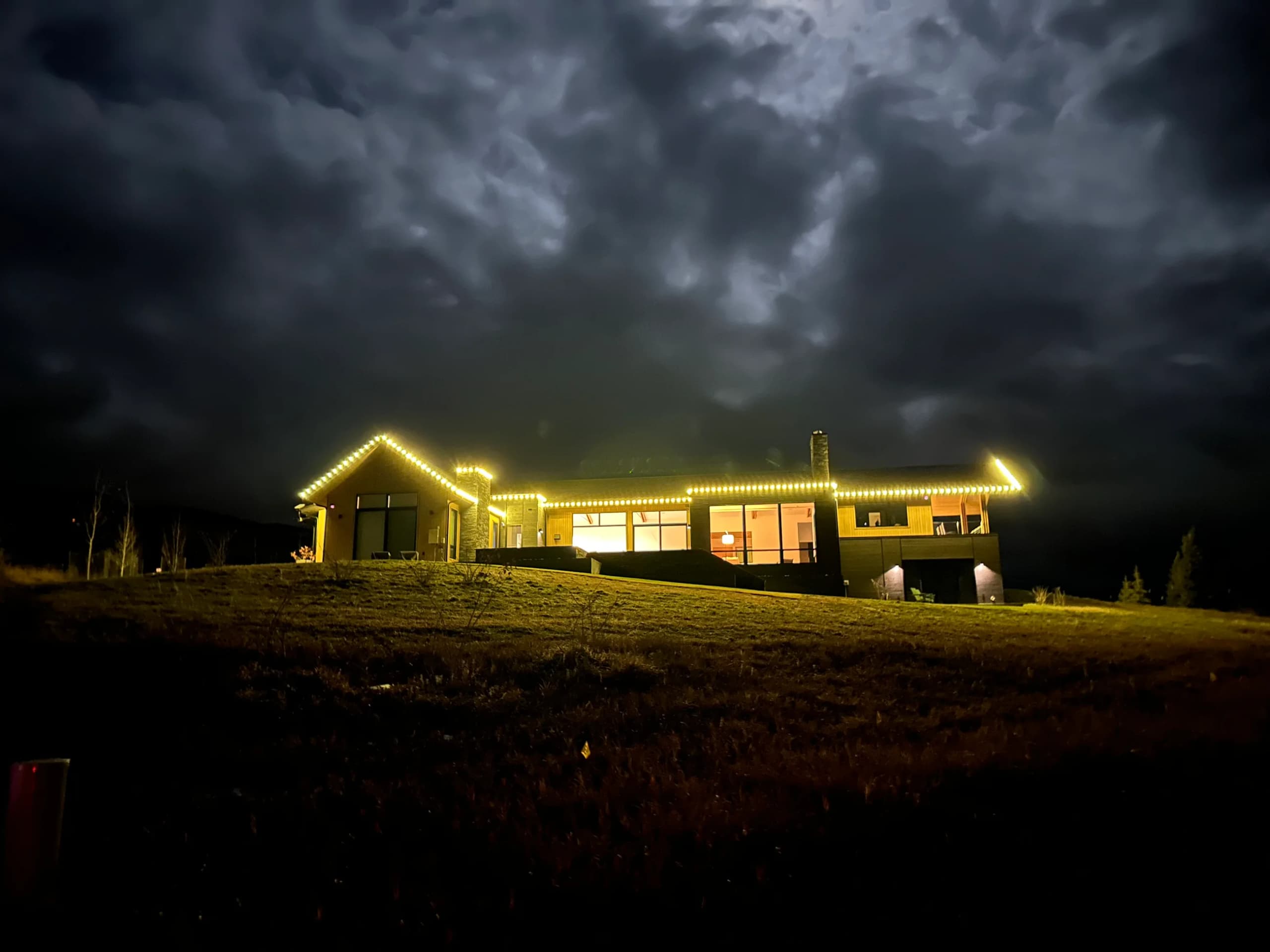 Single-story home outlined in gold roofline lights beneath dark storm clouds on open land