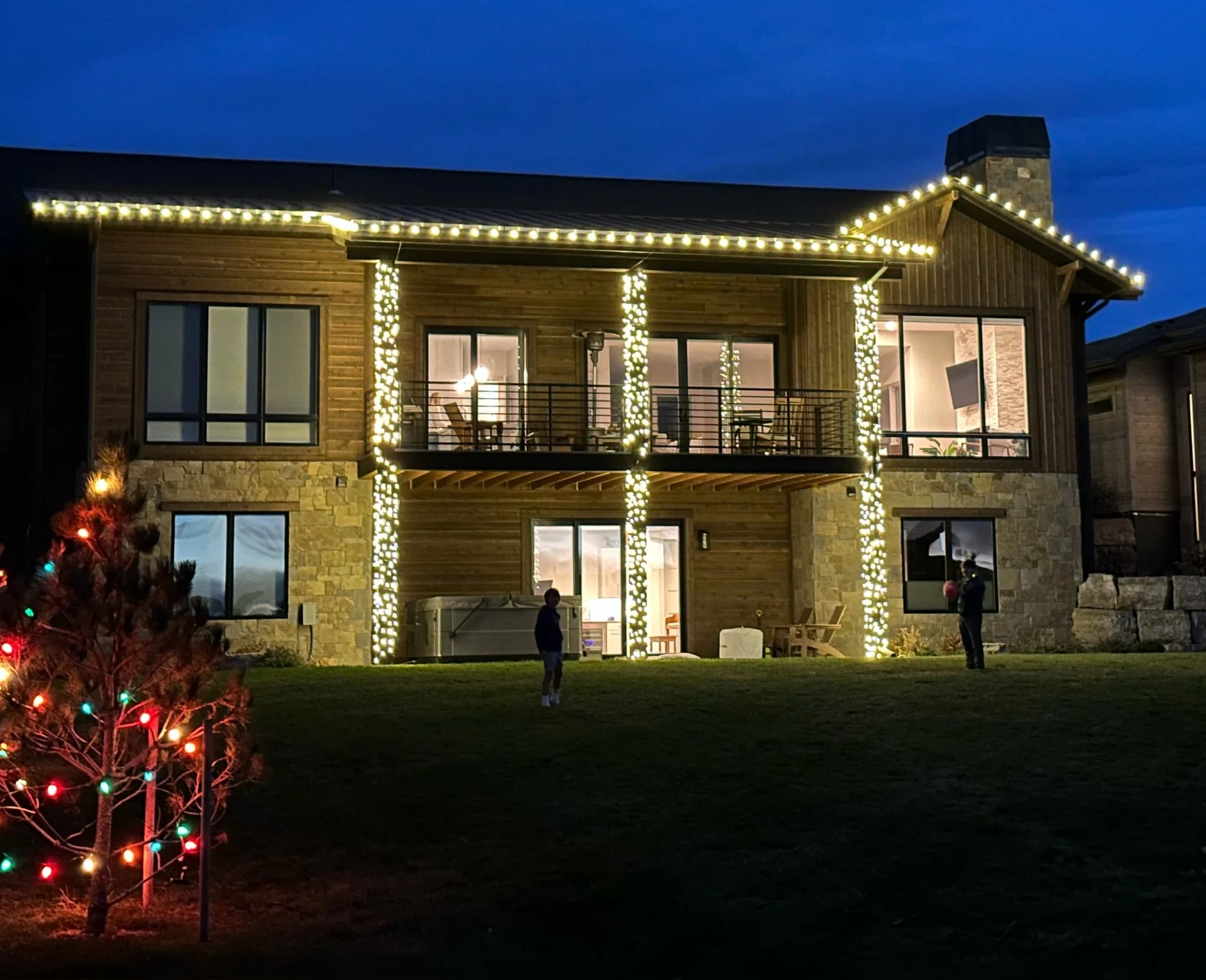 Two-story stone and timber home outlined in warm white roofline lights, with wrapped porch columns and a multicolor tree in the yard