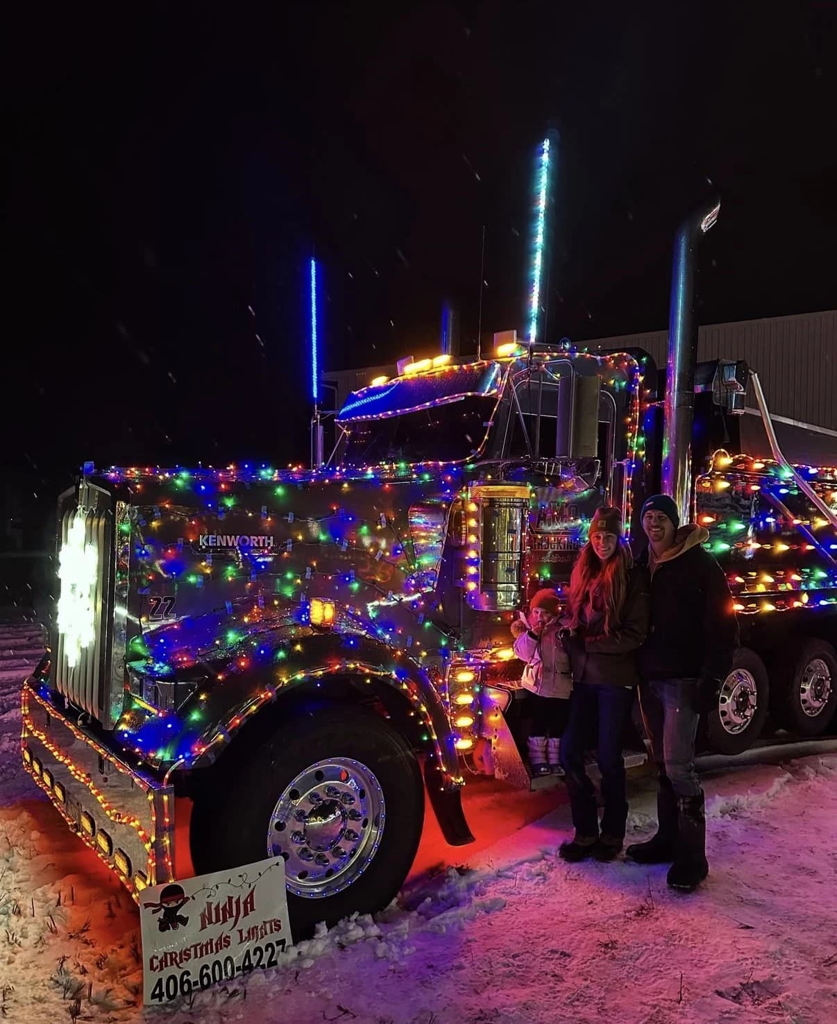 Tyler and Chelsea May with a young child, standing in the snow beside a semi truck covered in multicolor Christmas lights, with a Ninja Christmas Lights sign in front
